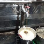 Finished maple syrup pouring from a stainless steel evaporator spout into a metal collection bucket.