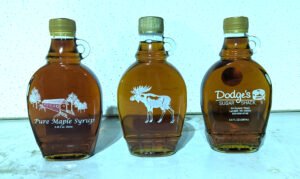 Three maple syrup glass bottles showing a covered bridge design, a moose design and the Dodge's Sugar Shack logo.