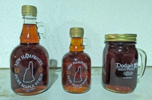 Two New Hampshire-labeled maple syrup glass bottles alongside a Dodge's Sugar Shack mason jar filled with dark amber syrup.