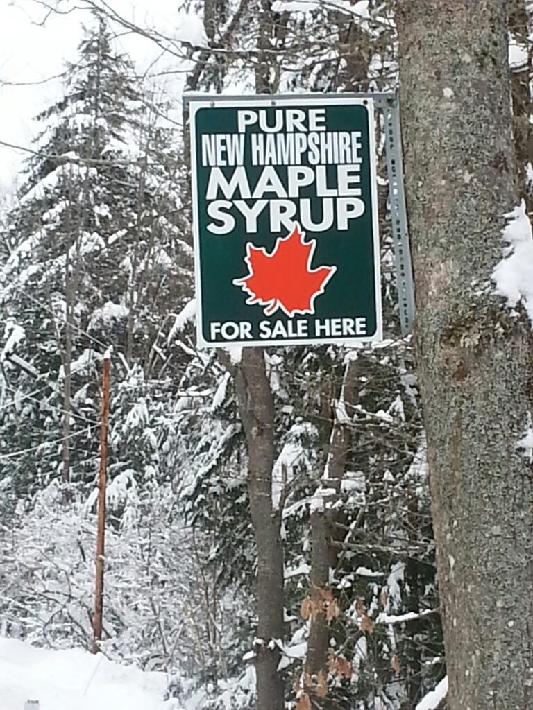 Roadside sign reads "Pure New Hampshire Maple Syrup For Sale Here" hanging on a snow-covered tree.